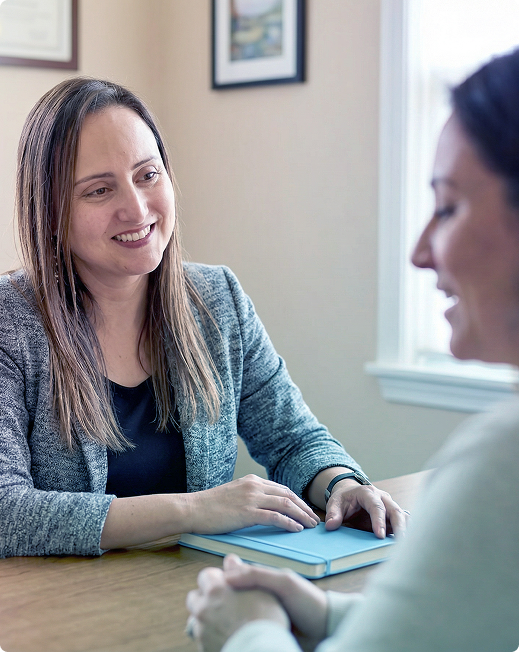 Two people talking at a table, one smiling with a notebook in a bright office.