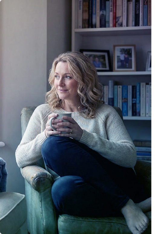 Woman sitting in a chair with a mug, by a bookshelf, looking out a window