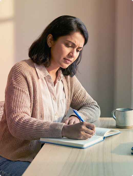 Person writing in a notebook at a table beside a mug, in soft natural light.