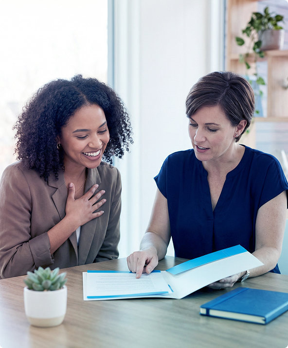 Two women reviewing documents at a desk with a blue binder and small potted plant in a bright office