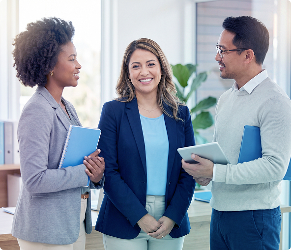Three coworkers chatting in a bright office, smiling and holding notebooks and tablets.