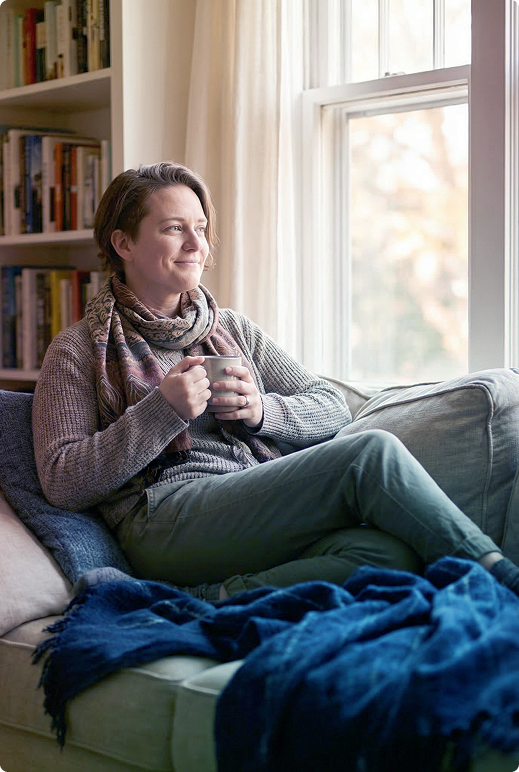 Person sitting on a couch by a window, holding a mug and looking out, with a blue blanket nearby.