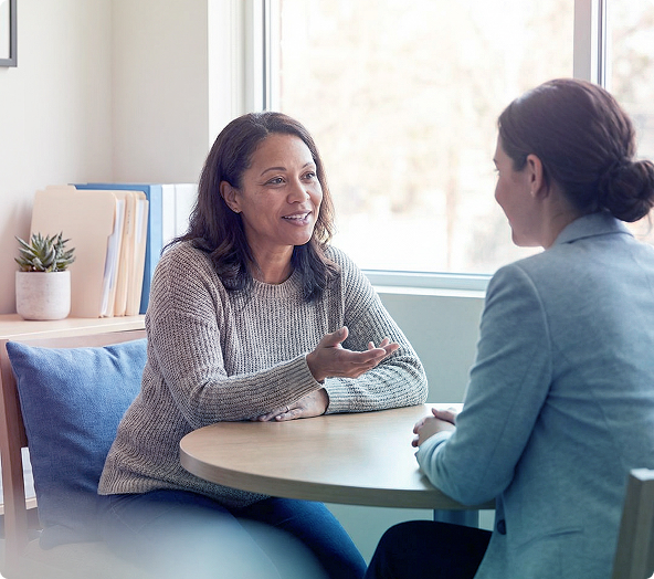 Two people talking at a round table in a bright office, one gesturing while seated on a blue couch.