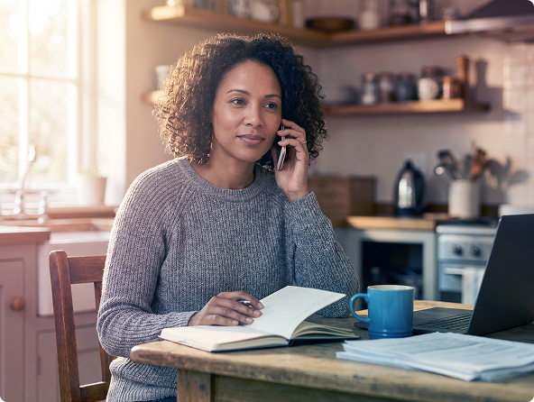 Woman reading a document at a desk beside a window, with a mug and blue notebook nearby.