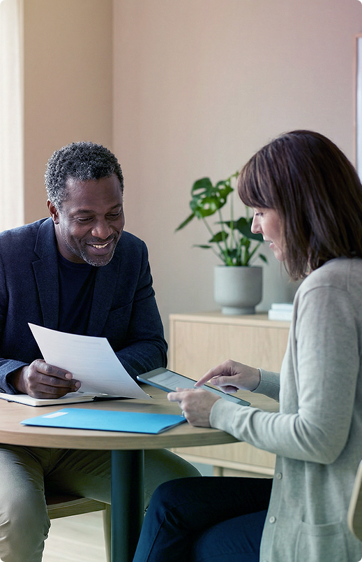 Two people discussing papers at a table in an office, with a plant in the background.