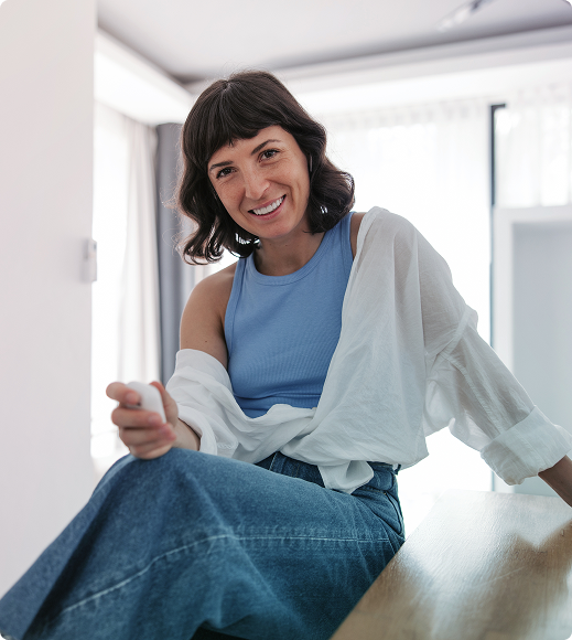 Smiling woman in a blue top and jeans sitting on a couch, holding a remote in a bright room