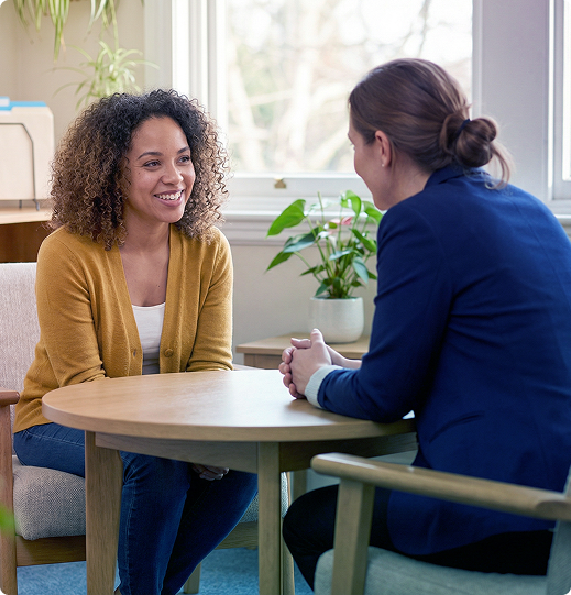 Two people talking at a round table in a bright office, one smiling and leaning forward.