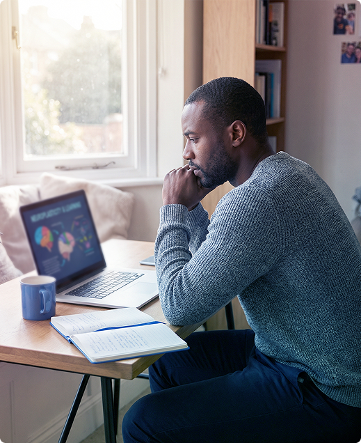 Person studying at a desk with a laptop, notebook, and mug by a sunlit window.