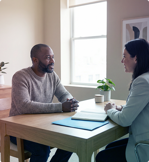 Two people in a bright office discuss papers across a wooden table by a window.
