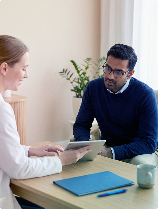 Two people in a meeting, one holding a tablet across a desk with papers, a notebook, and a mug.