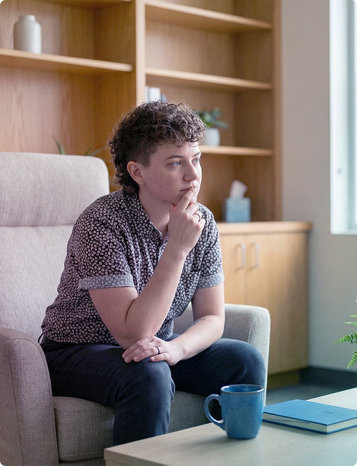 Person sitting on a chair, resting chin on hand, beside a blue mug in a cozy office lounge