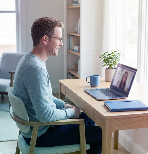Man on a video call at a desk with laptop, mug, and notebook in a bright home office