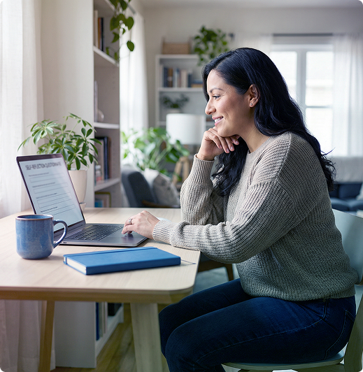 Woman working on a laptop at a desk in a bright home office, with a mug, notebook, and plants.