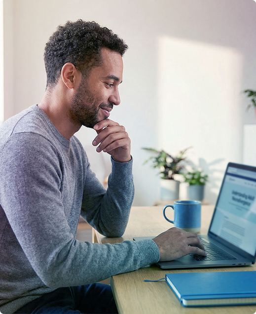 Person working at a desk on a laptop, resting chin on hand beside a blue mug and notebook.
