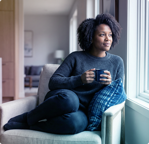 Person sitting by a window on a cushioned chair, holding a mug and gazing outside.