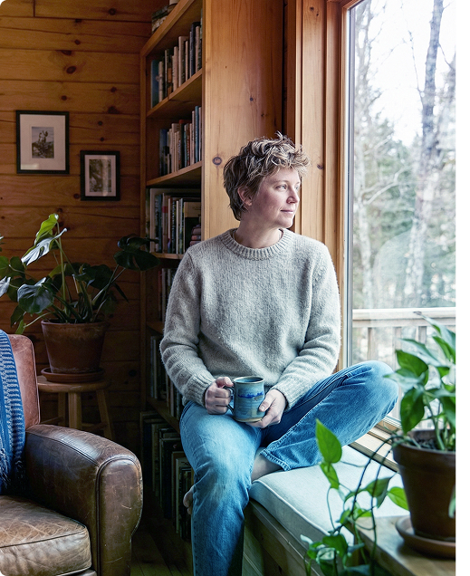 Person sits on a window seat holding a mug in a cozy wood-paneled room, looking outside.