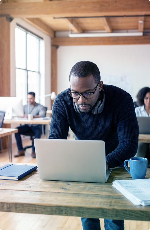 Person working on a laptop at a wooden table in a bright office, with a blue mug beside them