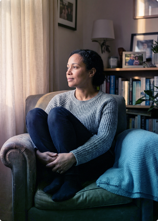 Woman sitting curled up in an armchair by a window, looking outside in a cozy living room.