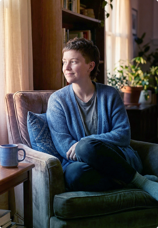 Person sitting in a green armchair by a window, looking out thoughtfully, with a blue mug on a nearby table.