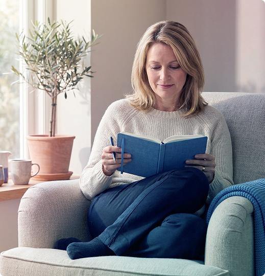Person reading a blue book on a gray sofa in a bright living room, with a potted plant nearby