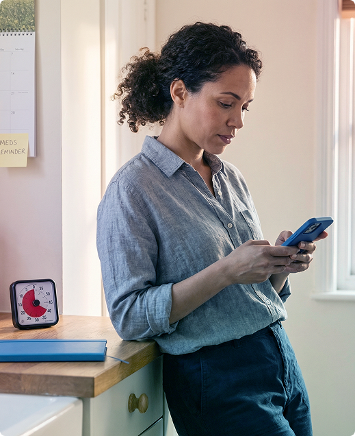 Person standing by a window, looking at a smartphone in a bright room.