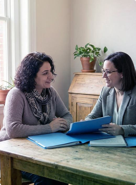 Two people talking across a table in a bright room, reviewing papers and a blue folder with a potted plant behind them