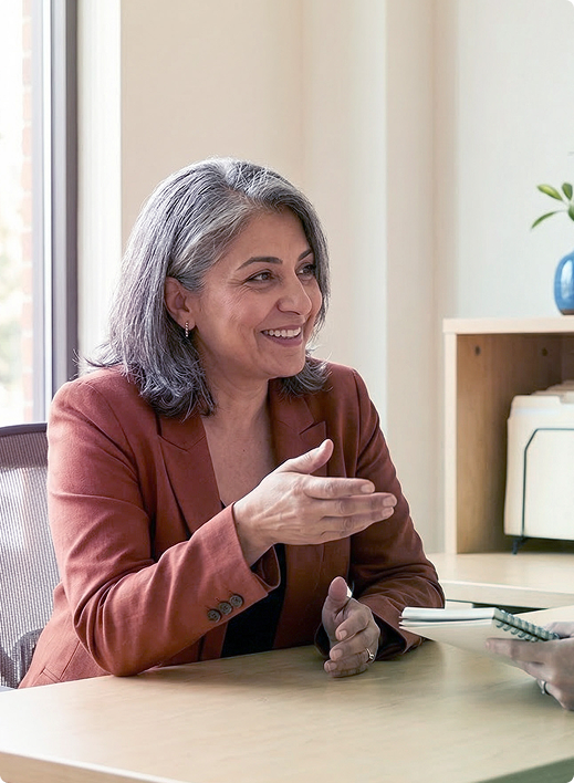 Woman in a rust blazer gesturing during a conversation at a table in a bright office