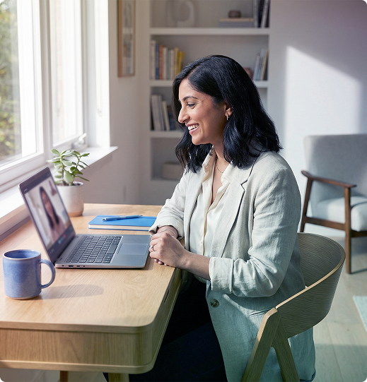 Woman smiling while video chatting on a laptop at a sunlit desk with a coffee mug