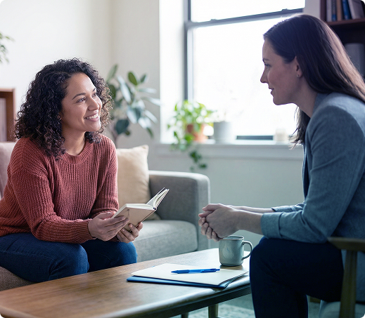Two women talk on a couch in a bright living room, one holding papers and the other a mug.