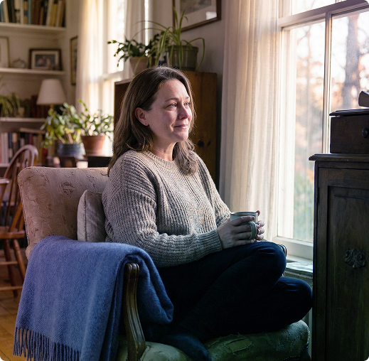 Woman sitting in an armchair by a sunlit window, wrapped in a blanket, smiling toward a stove.