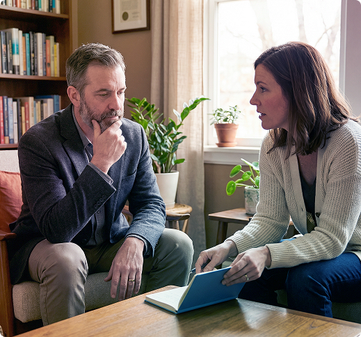 Two people talking in a cozy living room, with one holding a notebook and the other listening thoughtfully.