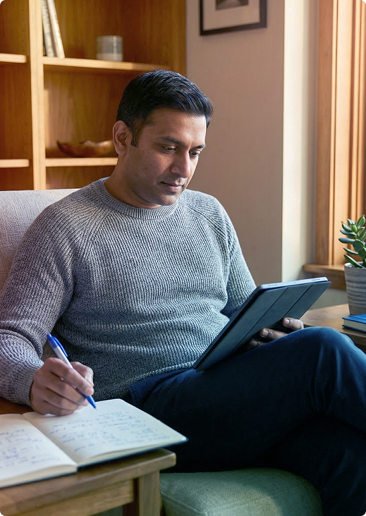 Person sitting on a couch, writing in a notebook while using a tablet in a cozy room.