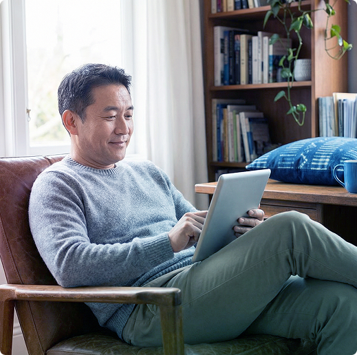 Man sitting on a chair using a tablet in a bright living room beside a bookshelf.