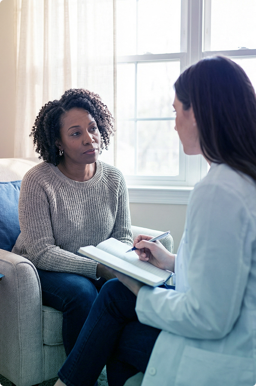Two women talking during a counseling session, one taking notes on a clipboard near a bright window.
