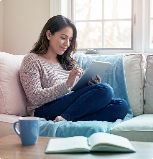 Woman writing on a tablet while sitting on a couch by a window, with an open book and mug nearby