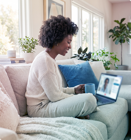 Person sitting on a sofa using a laptop and holding a mug in a bright living room