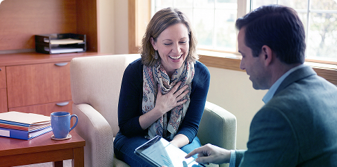 Two people talking on a couch in a bright office, one holding a clipboard and smiling