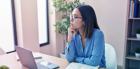 Woman in blue shirt working at a laptop, seated at a desk in a bright office.