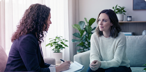 Two women talking in a living room, one holding a notebook, with plants and shelves in the background