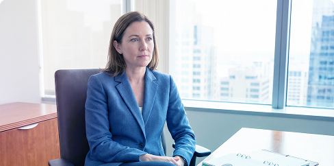 Woman in a blue blazer seated by a bright office window, looking ahead with papers on the desk.