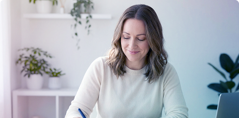 Woman writing at a desk in a bright home office with plants and a laptop