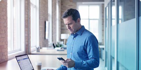 Man in blue shirt using a laptop and smartphone in a bright office hallway