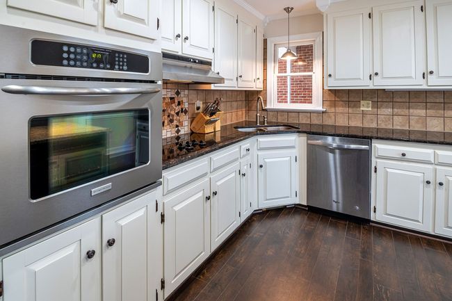 Bright white kitchen with stainless steel oven and dishwasher, dark countertops, and dark wood floor.