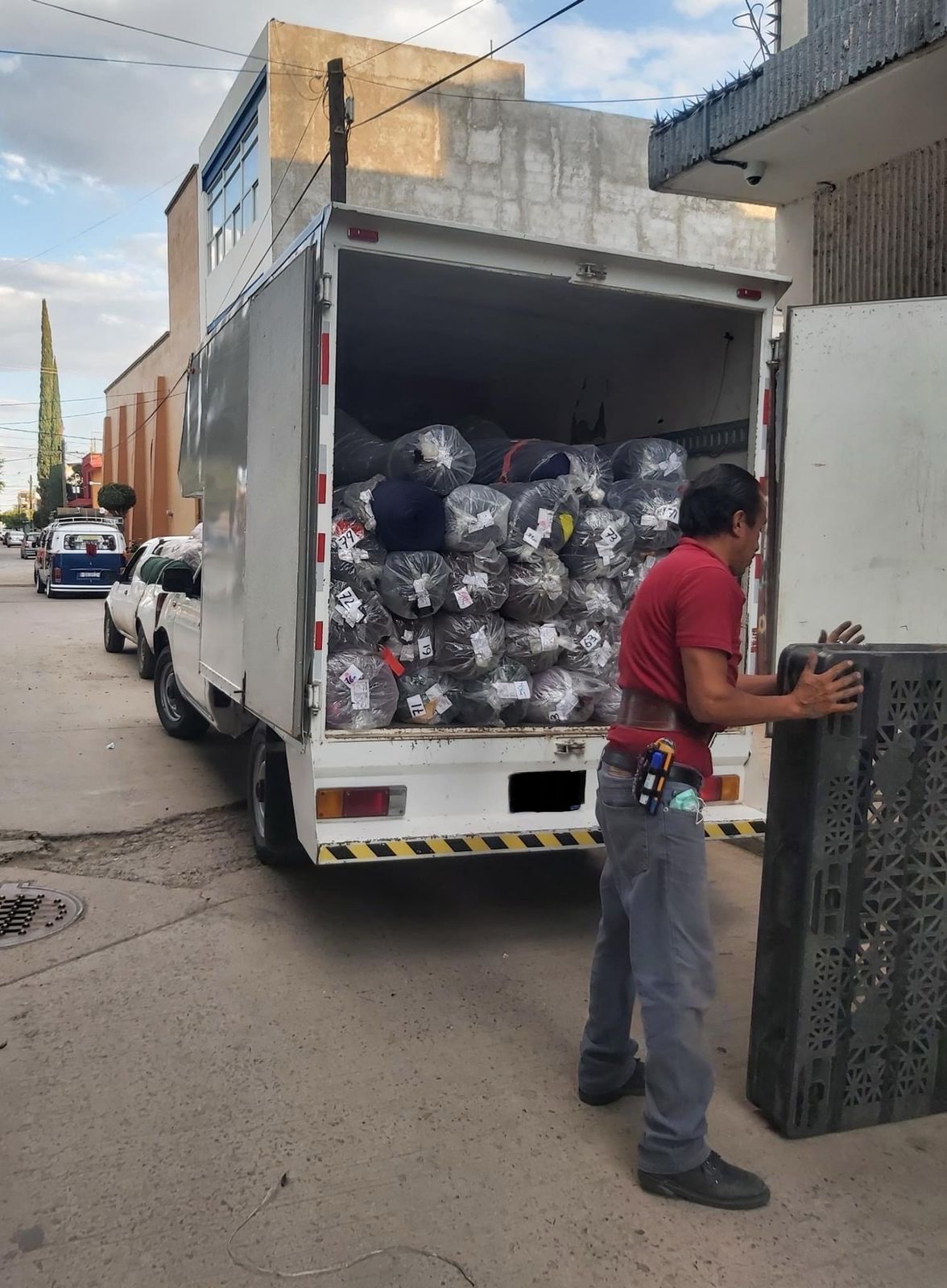 Un hombre con una camisa roja está parado junto a un camión lleno de bolsas de ropa.