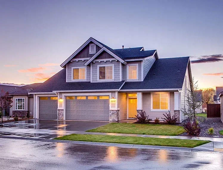 Two-story suburban house with a gray exterior, three-car garage, and wet driveway at dusk.
