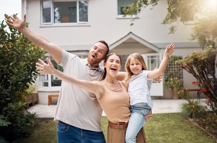 Family of three happily embracing in front of their house, arms raised.