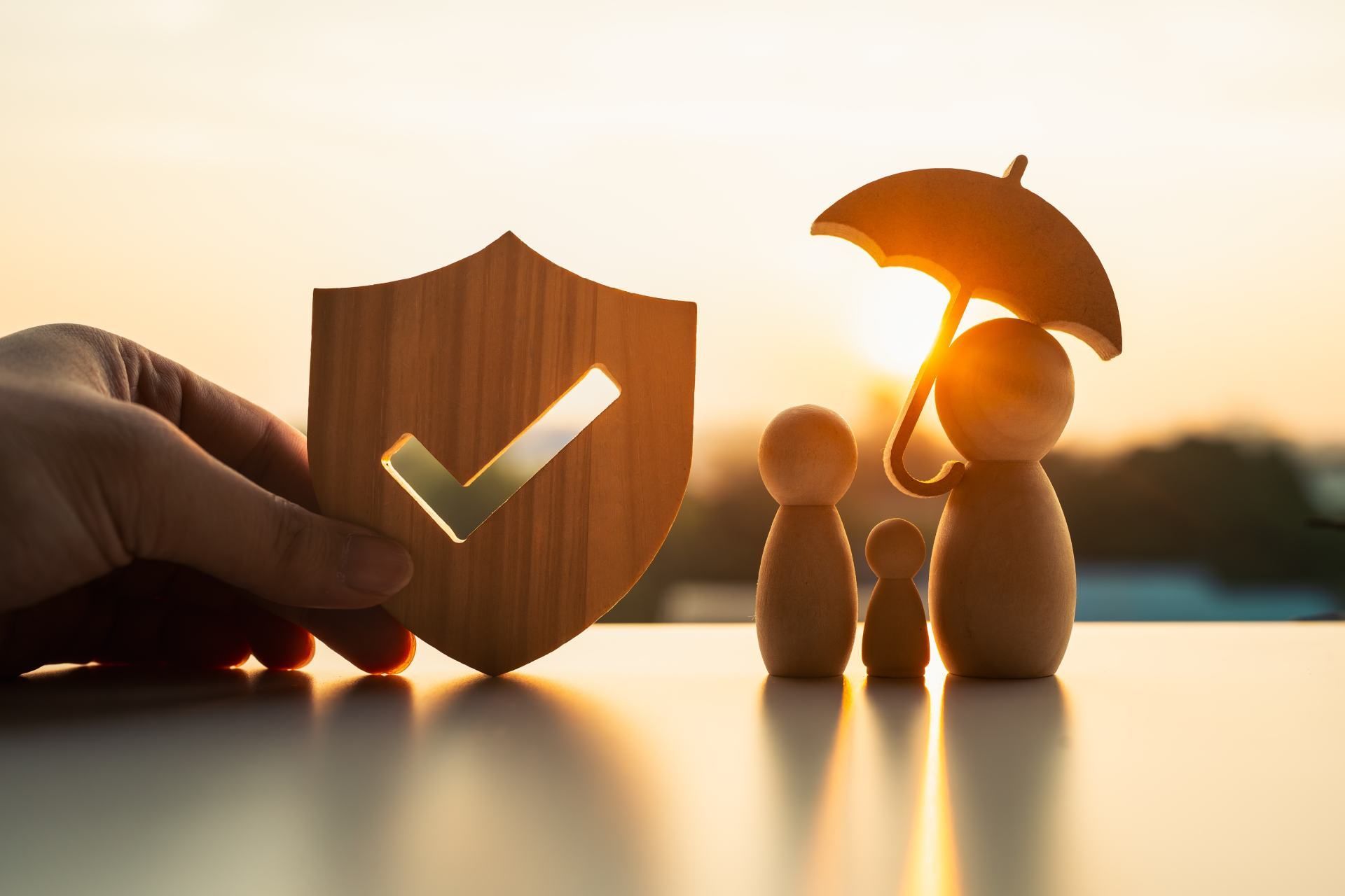 Hand holding a wooden shield with a checkmark next to a family figurine under an umbrella, set against a sunset.