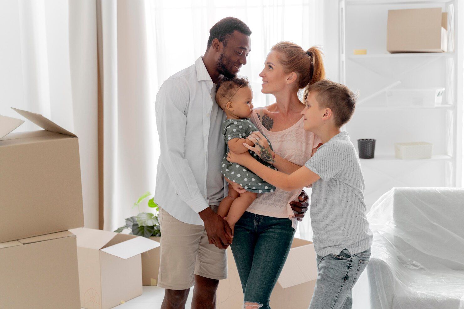 Family of four in a new home, boxes around them. Parents holding a baby, son hugs mom.