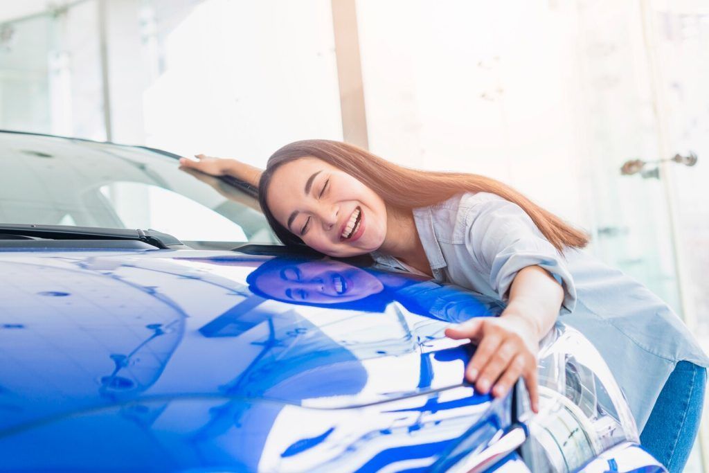 Smiling woman hugging the hood of a blue car, happy expression. Bright interior setting.