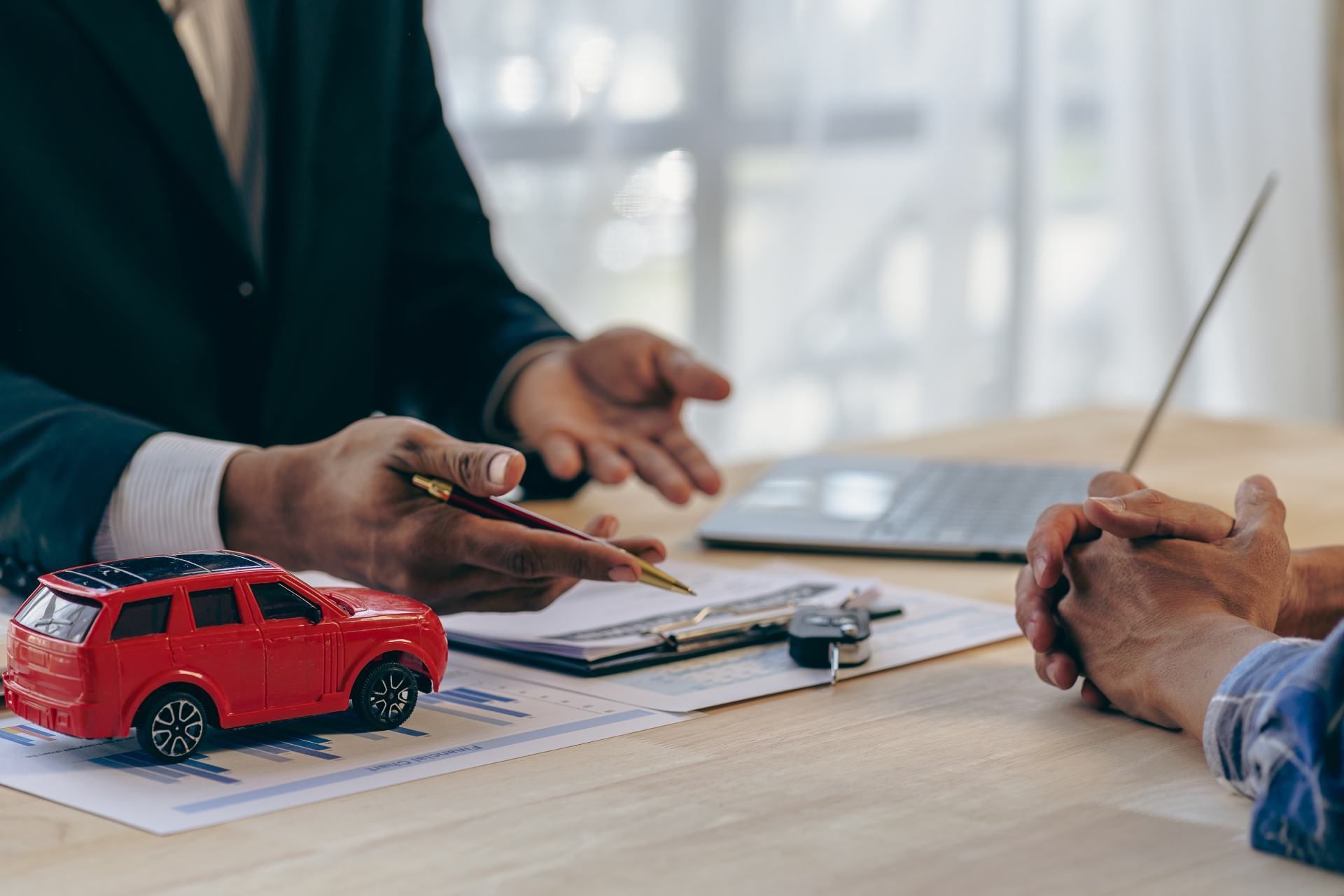 Person in suit points at paperwork as a client listens, a red toy car sits nearby on a table.
